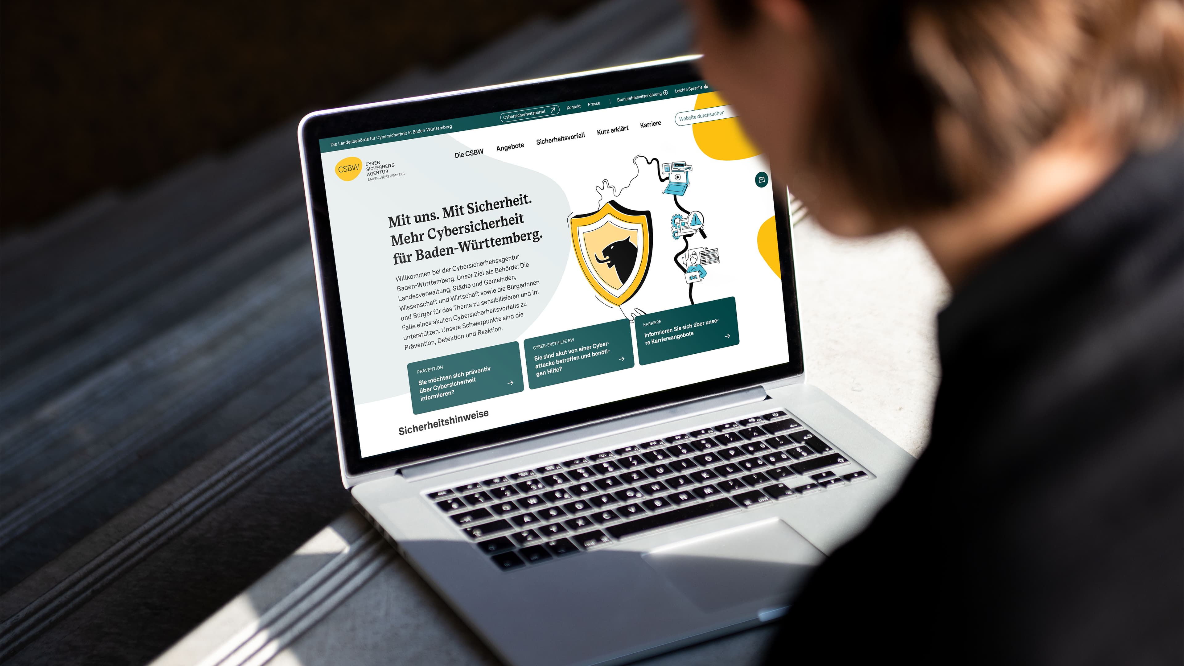 Eine Person betrachtet auf einem Laptop die Website der Cybersicherheitsagentur Baden-Württemberg. Auf dem Bildschirm sind ein Schutzschild mit Panther-Symbol sowie Informationen und Angebote rund um Cybersicherheit zu sehen.
